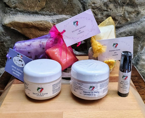 Una selezione di prodotti per la cura della pelle a base di lavanda dell’azienda agricola Torre Memoriola è disposta su un vassoio di legno, su sfondo di pietra rustica. Il set include barattoli e confezioni con etichette in italiano, come shampoo solido, balsamo solido, saponetta alla lavanda, crema al burro di karité, sapone da barba e olio roll-on, tutti in contenitori eco-sostenibili.